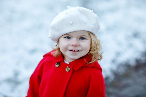 Outdoor winter portrait of little cute toddler girl in red coat and white fashion hat barret. Healthy happy baby child walking in the park on cold day with snow and snowfall. Stylish clothes for kids