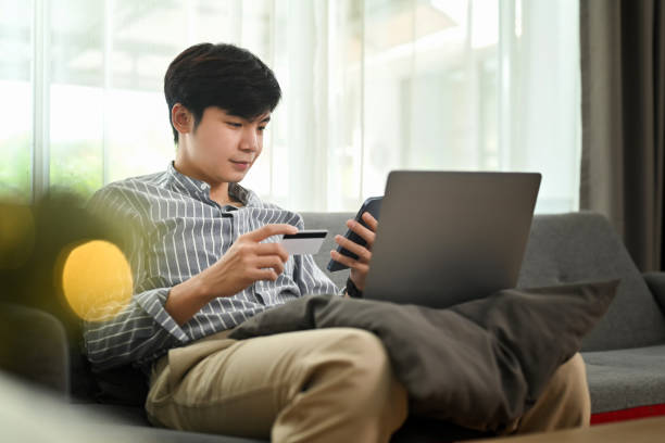 Satisfied millennial man holding credit cad and mobile phone, entering data for website form. E-commerce concept. Satisfied millennial man holding credit cad and mobile phone, entering data for website form. E-commerce concept. asian man online shopping stock pictures, royalty-free photos & images