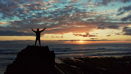 Human with raised hands looks to the sun over horizon in morning while sunrise.  Man stands on a rock against the ocean  beautiful summer sunset. Beauty of nature.  Silhouette of a man against the sky