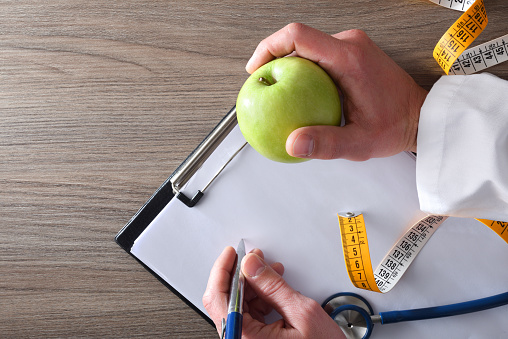 Nutritionist doctor taking notes in his notebook with apple in hand for body check with stethoscope and meter on wooden table. Top view.