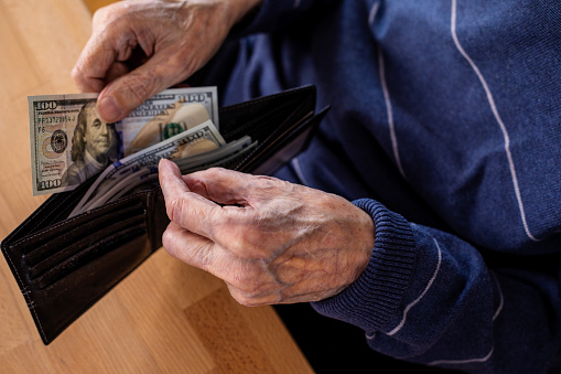 Elderly man holding wallet and counting money, close-up of wrinkled human hands holding US paper currency