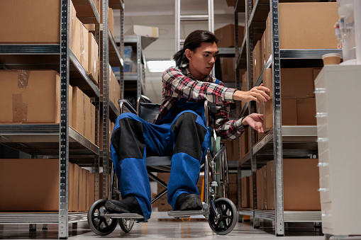 Warehouse asian package handler in wheelchair searching cardboard box on shelf. Shipment manager with disability taking parcel from rack while working in inclusive workplace