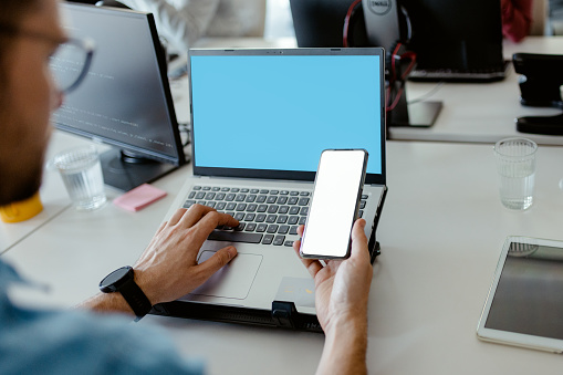 Male hands holding smartphone with empty white blank screen mockup while working at desk in office, it developer concept, copy space