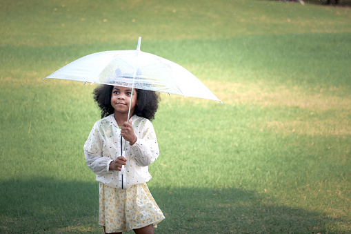 Happy smiling African girl with black curly hair with umbrella under raindrops fall while standing outdoor green park, beautiful kid playing outside garden on rainy day, cute child playing in the rain