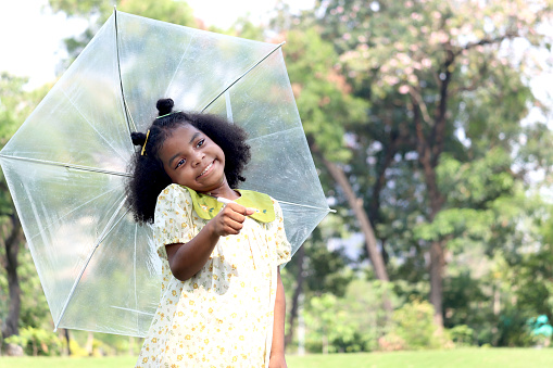 Happy smiling African girl with black curly hair with umbrella under raindrops fall while standing outdoor green park, beautiful kid playing outside garden on rainy day, cute child playing in the rain