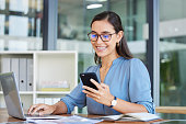 Business woman, laptop and smile for phone, communication or reading at the office. Happy female employee event planner checking email, social media or post on smartphone and computer at workplace