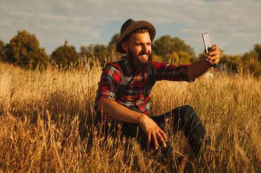 Glad bearded man in checkered shirt and hat smiling and taking selfie via cellphone while sitting on grass on autumn day in meadow