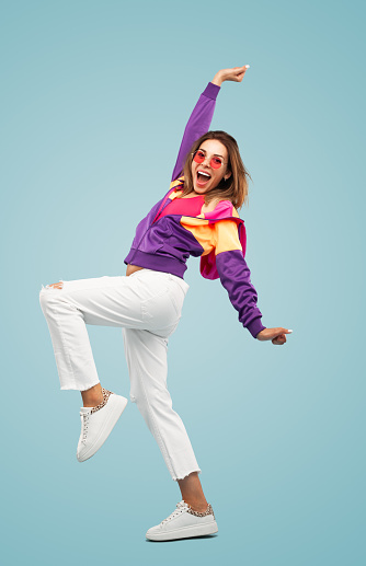 Side view of excited young female millennial in stylish outfit and sunglasses screaming happily and looking at camera while standing against blue background with raised arm