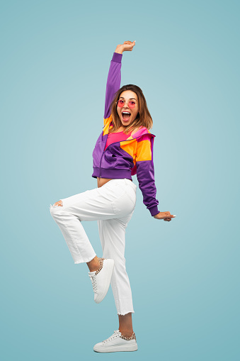 Full body amazed young female in colorful clothes with sunglasses raising arm and leg and yelling in excitement after victory against blue background