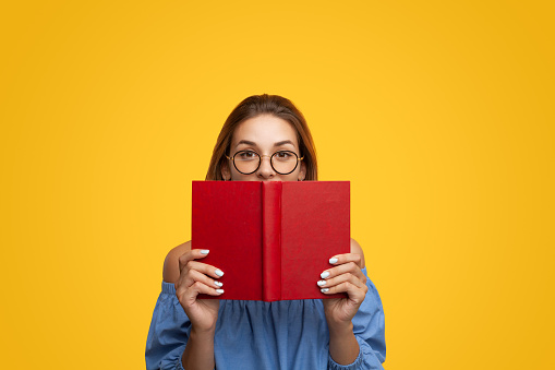 Smart young female with long brown hair in blouse and eyeglasses covering mouth with red book and looking at camera against yellow background