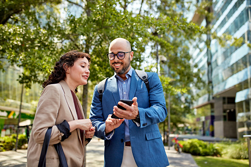 Waist-up view of mid adult professionals standing outdoors in sunny springtime weather and looking at photos on portable device.