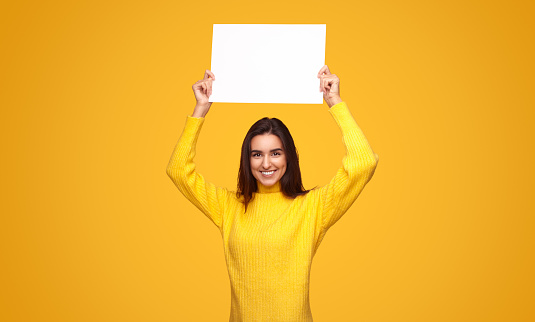 Positive young ethnic female with long dark hair in casual clothes smiling and looking at camera while demonstrating blank banner against yellow background