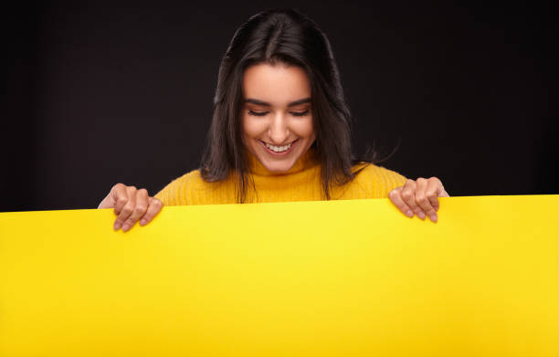 istock Happy ethnic lady smiling while showing yellow poster in black studio 1475804385
