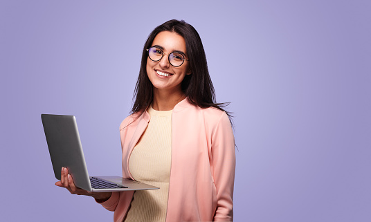 Positive young ethnic female freelancer with long dark hair in trendy outfit and eyeglasses smiling and looking at camera while working remotely on laptop against violet background
