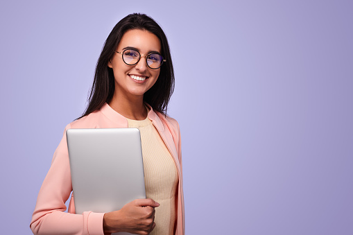 Confident young ethnic female student with long dark hair in eyeglasses smiling and looking at camera while standing against purple background with laptop