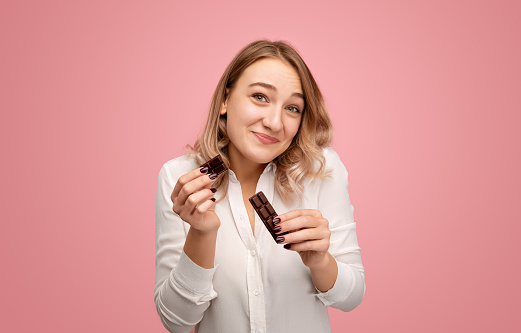 Young female in white blouse shrugging shoulders and looking at camera with apologetic smile while breaking chocolate bar against pink background
