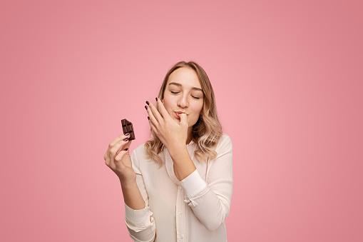 Young female in white blouse closing eyes and licking thumb while eating tasty chocolate against pink background