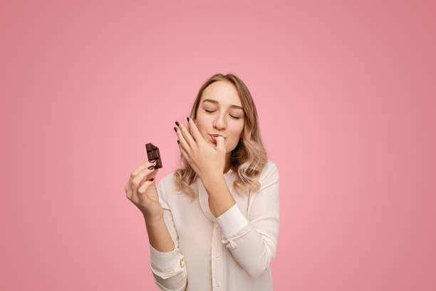 Young woman with chocolate licking finger Young female in white blouse closing eyes and licking thumb while eating tasty chocolate against pink background chocolate fingers stock pictures, royalty-free photos & images