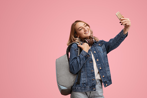 Cheerful young female student with long hair in casual clothes backpack and headphones on neck smiling while taking selfie on smartphone against pink background