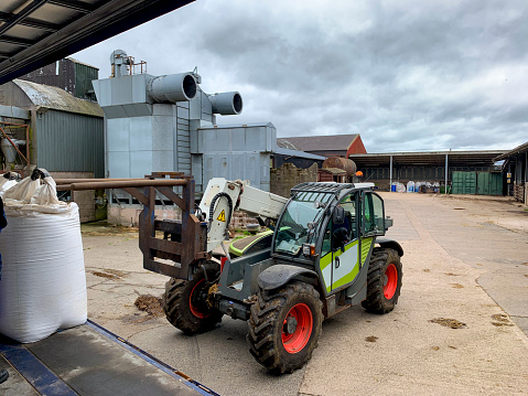 A stationary telescopic handler on a farm in Embleton, North East England. There are agricultural buildings in the background of the shot and the tele handler is ready to pick up bags of seeds.