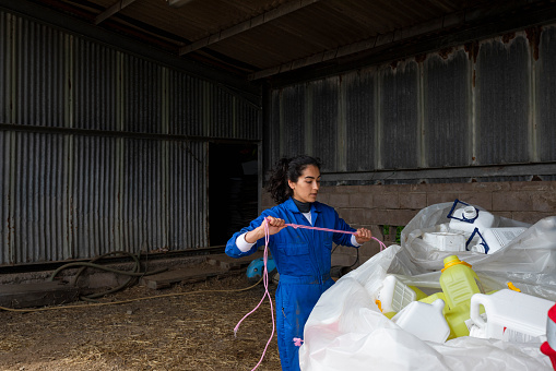 A young female farmer wearing overalls while completing her daily tasks at the farm where she works in Embleton, North East England. She is tying up plastic bags filled with empty manufactured fertiliser bottles with rope, ready to take them to the recycling bank.