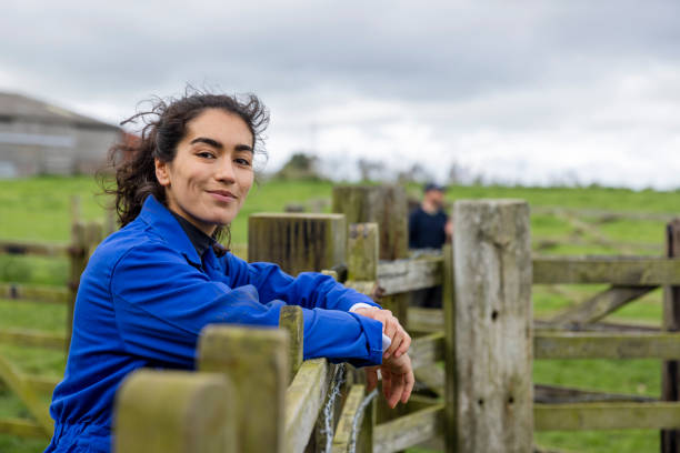 Confident in Her Line of Work A portrait of a young female farmer wearing overalls, standing and leaning against a fence at the farm she works at in Embleton, North East England while looking at the camera and smiling. Farmer stock pictures, royalty-free photos & images