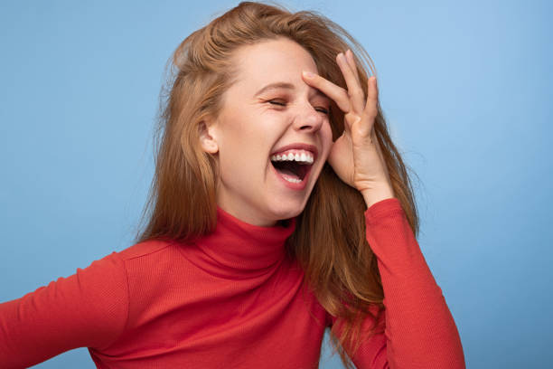 Cheerful redhead woman laughing at joke Happy young female in red turtleneck with ginger hair touching forehead and laughing with closed eyes against blue background people laughing hard stock pictures, royalty-free photos & images