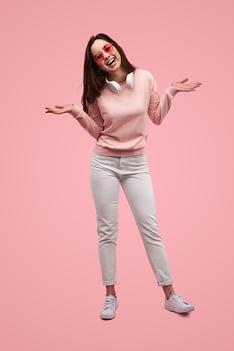 Full body of young cheerful female model in casual outfit and sunglasses shrugging shoulders and laughing while standing against pink background