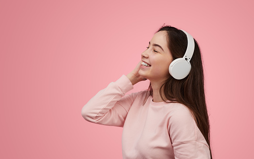 Cheerful teen meloman in sweatshirt touching wireless headphones and smiling with closed eyes while enjoying good music against pink background