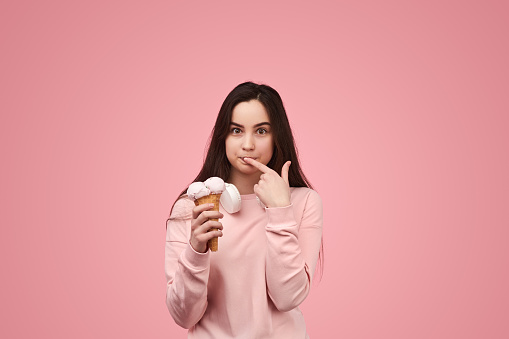 Hungry young female millennial with long dark hair in casual clothes and headphones on neck touching lips with finger and looking at camera with excitement while eating delicious ice cream