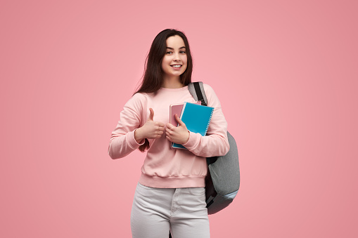 Happy teenager with backpack and notebooks looking at camera with smile and gesturing thumb up while approving studies against pink background