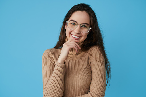 Clever teen student in turtleneck and glasses looking at camera with smile and touching chin while standing against blue background