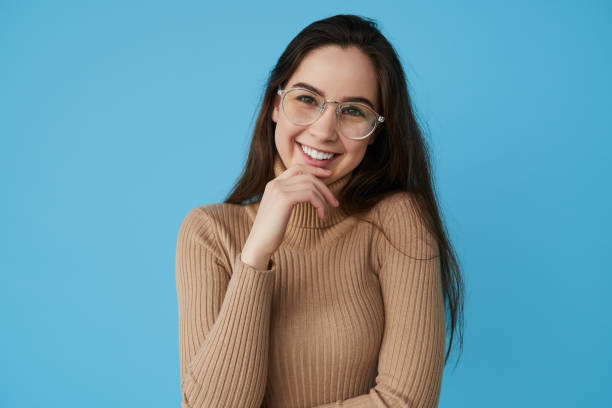 Smart teenager touching chin and smiling Clever teen student in turtleneck and glasses looking at camera with smile and touching chin while standing against blue background nerd sweater stock pictures, royalty-free photos & images