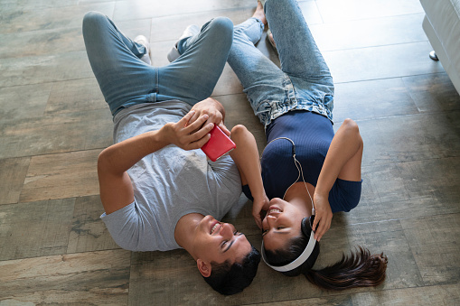 Happy and young couple in love listening to music from a smart phone resting on their home's floor. Overhead shot.