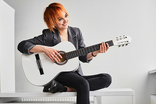 Happy ginger female musician in casual clothes smiling and looking away while sitting on shelf and playing acoustic guitar during lesson in light classroom
