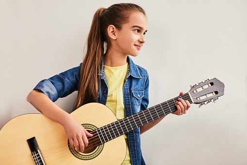Positive teenager in casual clothes with ponytail playing acoustic guitar and looking away during lesson against gray wall