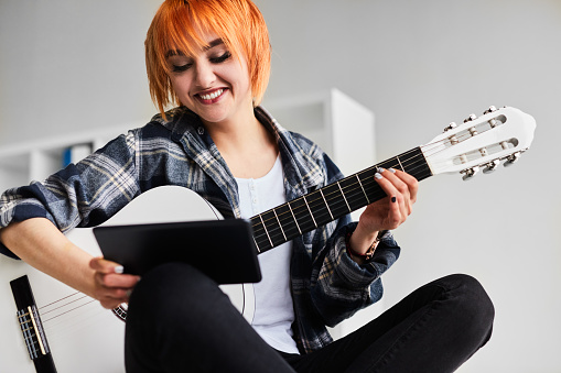 Crop positive young female musician with short dyed red hair and bright makeup in checkered shirt smiling and watching tutorial video on tablet while playing acoustic guitar in white room