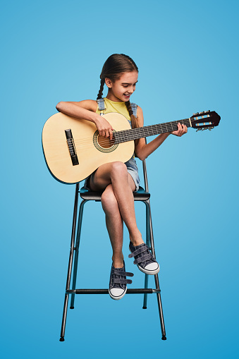 Full body teenager in casual clothes smiling and playing acoustic guitar while sitting on chair during music lesson against bright blue background