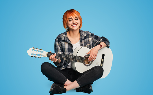 Gleeful redhead female musician in checkered clothes looking at camera with smile and playing white acoustic guitar while sitting cross legged against blue background