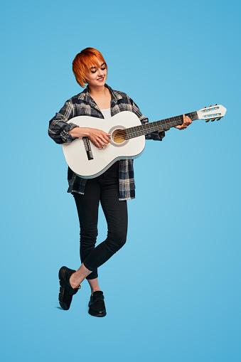 Full body glad female musician in casual clothes with short ginger hair smiling and playing acoustic guitar against blue background