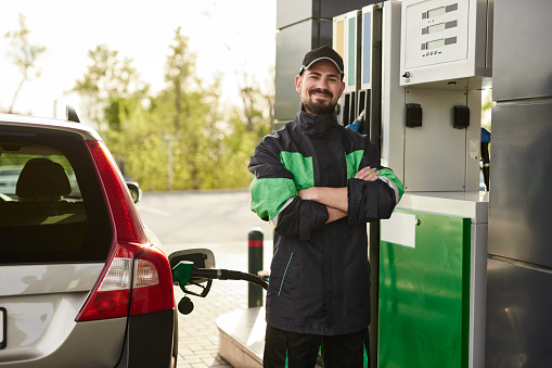 Confident bearded man in uniform crossing arms and looking at camera with smile while standing between vehicle and pump on sunny day at gas station