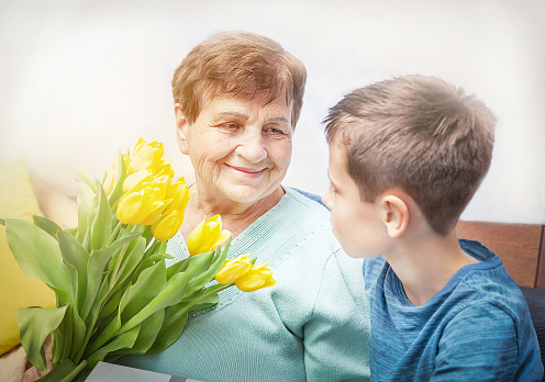 Boy giving a bunch of yellow flowers to grandmother. Grandson and grandma spending, enjoying time together.  Kindness to senior (elderly) woman. Happy Mother's Day, grandmother day, International Women's Day.