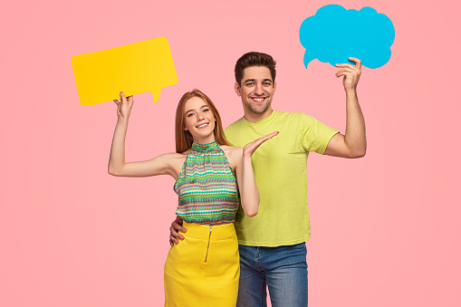 Excited young couple in casual clothes hugging and smiling at camera while demonstrating blue and yellow empty speech bubbles against pink background