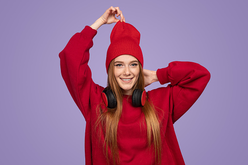 Happy young redhead woman in red sweatshirt with wireless headphones looking at camera with smile and adjusting beanie against violet background