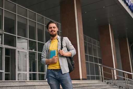Low angle of positive young bearded male student in casual clothes with backpack and notebook smiling and looking at camera while standing on stairs of university building