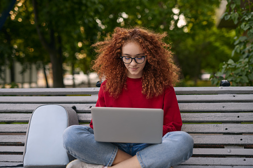 Glad young woman in casual clothes and glasses with curly red hair sitting cross legged on bench near backpack and browsing netbook while studying in park in summer