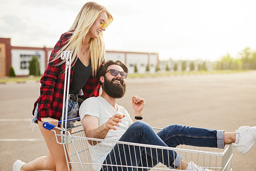 Cheerful young woman in casual clothes with long blond hair smiling and pushing shopping trolley with bearded boyfriend while having fun on parking lot outside supermarket
