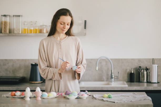 istock Young woman decorating Easter eggs using brush and paint on the kitchen. 1475819485