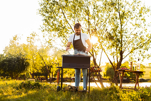 Full body male chef in apron smiling and grilling sausages near tree on grassy shore of calm lake on sunny weekend day