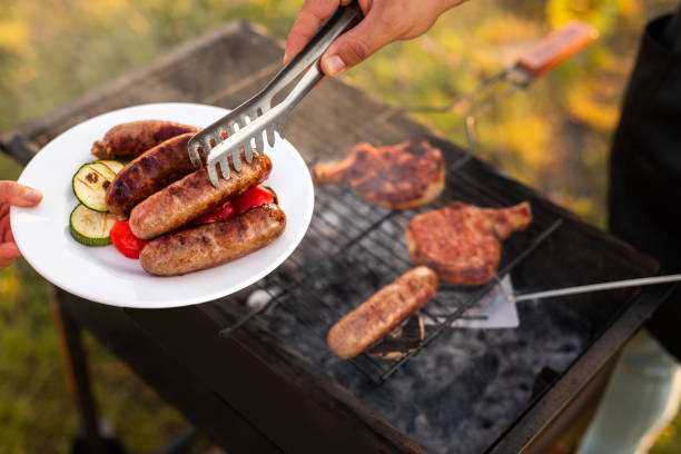 Crop chef serving sausages and vegetables during picnic stock photo